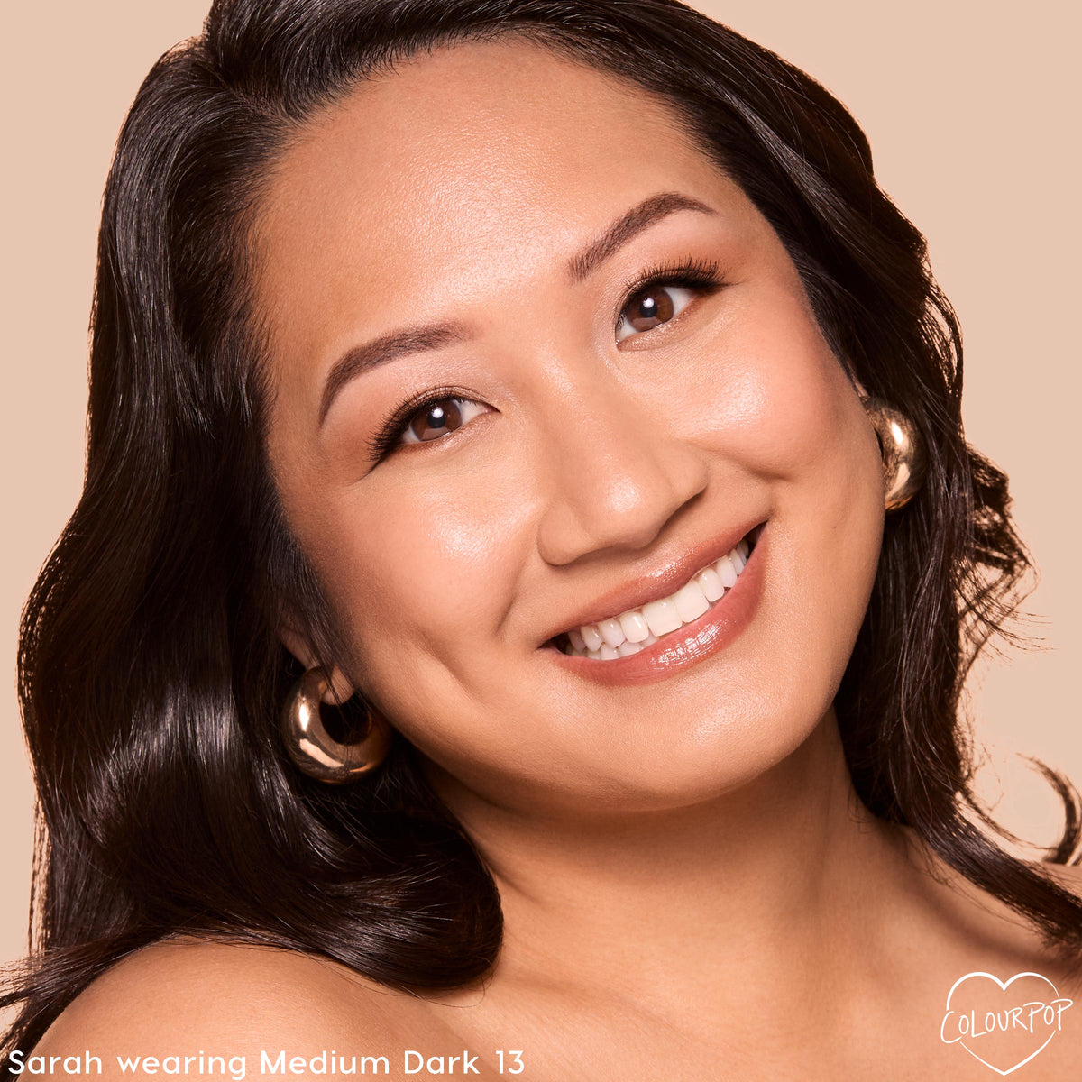 Model smiles wearing ColourPop Pretty Fresh Tinted Foundation Balm in Medium Dark 13, showcasing dewy, even coverage on a close-up face with soft glam eye makeup and gold hoop earrings against a warm beige studio background.