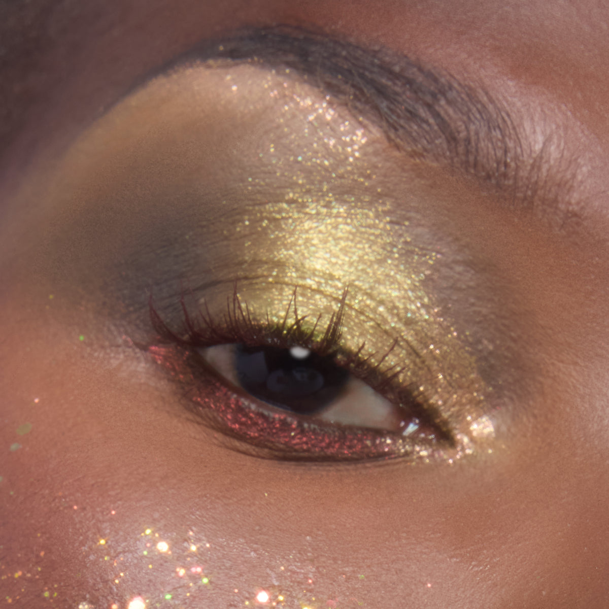 Close-up eye wears metallic lime-gold sparkle on the lid and red shimmer along the lower lashes, showcasing ColourPop Glitter Gel in Ogres are Like Onions, with chunky glitter flecks dusted across the cheekbone.
