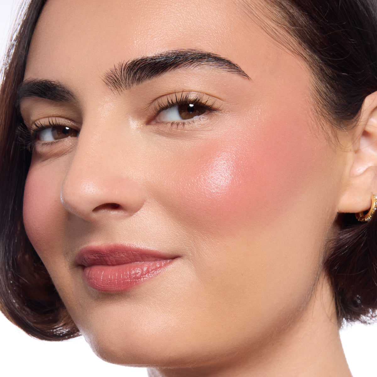 Model's cheeks and lips wear ColourPop Lip and Cheek Balm, Fairytale Things, adding a dewy rosy flush to cheeks and a soft lip tint in a studio close-up, highlighting smooth skin, natural brows, and a small gold hoop earring.