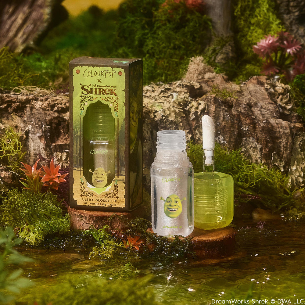 ColourPop Ultra Glossy Lip in Ogre, open with applicator upright beside its box; frosted tube with Shrek face and green cube cap holds a clear, shimmery gloss, staged on a wood slice by water amid moss and flowers in a swamp setting.