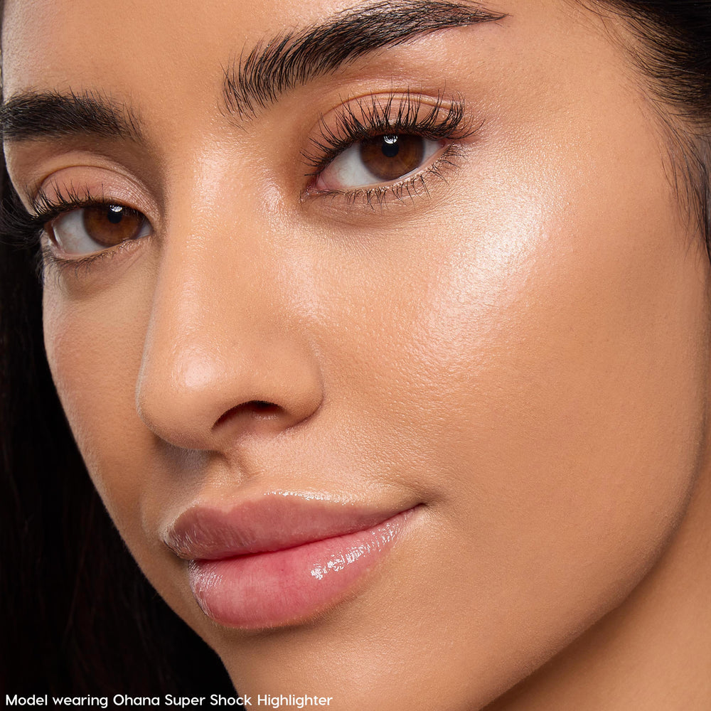 Model wearing Ohana ColourPop Super Shock Highlighter catching light on the cheekbone, delivering a pearly champagne glow with a smooth, dewy finish; close-up beauty shot shows natural skin texture, soft lighting, glossy lips, and defined brows.