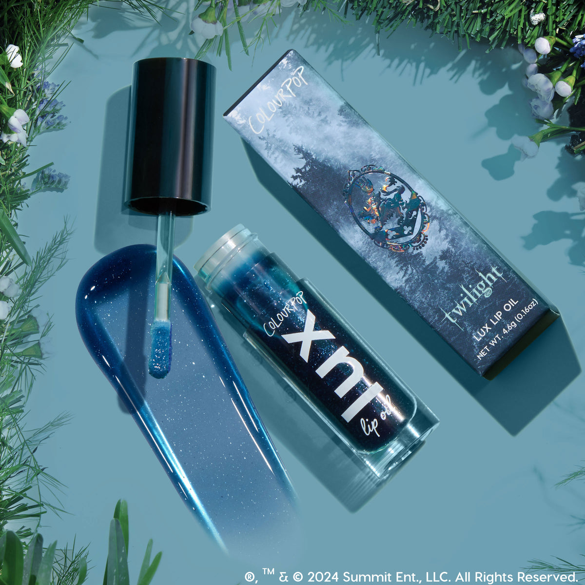 Team Edward Lux Lip Oil swatched: a sparkling deep blue, shimmer-packed gloss. Doe-foot applicator, open ColourPop Lux Lip Oil tube, and Twilight collab box rest on a teal surface framed by evergreen sprigs.