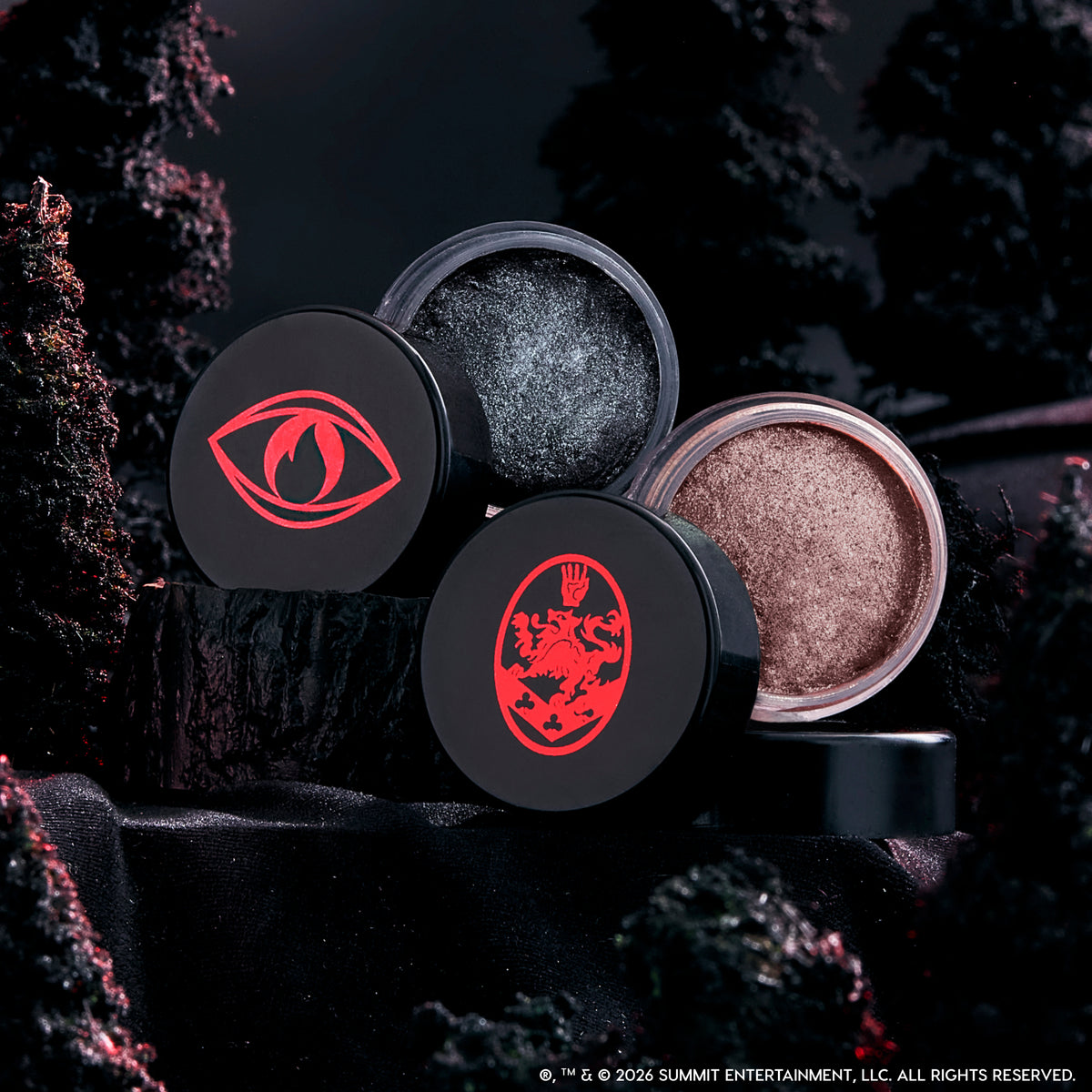 Coven Clash Jelly Much Duo eyeshadows sit open on black fabric, revealing shimmery charcoal and champagne shades in round pots with bold red Twilight-inspired symbols, styled against a dark, moody forest backdrop.