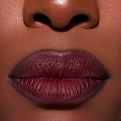 Close-up of full lips wearing Newborn powder to creme lipstick in a deep twilight berry shade, showing smooth, rich pigment and velvety matte-satin finish on dark skin for a bold, dramatic lip look.