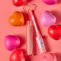 ColourPop Ultra Glossy Lip in One Kiss, a peachy‑pink shimmer, shown with open tube and applicator creating a heart-shaped swipe; pink cap with iridescent hearts; surrounded by red, pink, and orange heart props on a coral background.