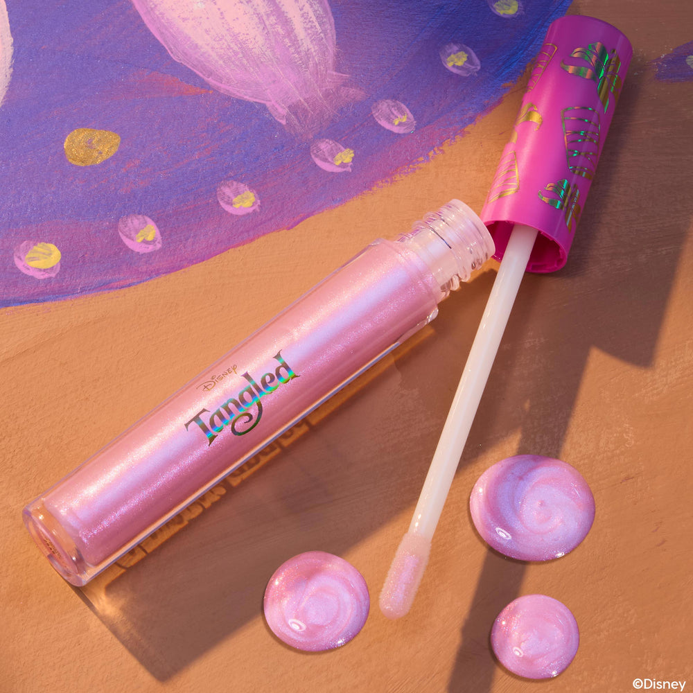 Rapunzel ColourPop Ultra Glossy Lip, an iridescent pink shimmer gloss, lies open with a doe-foot applicator and glossy swirls, featuring a hot-pink lantern-print cap, styled on a tan surface beside purple Tangled artwork from the Disney collab.