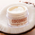 Open jar of Coconut overnight lip mask showing a swirl of creamy balm, resting on a halved coconut; clear pot with tan lettering highlights hydrating formula for dry lips; ColourPop Lip Mask product shot.