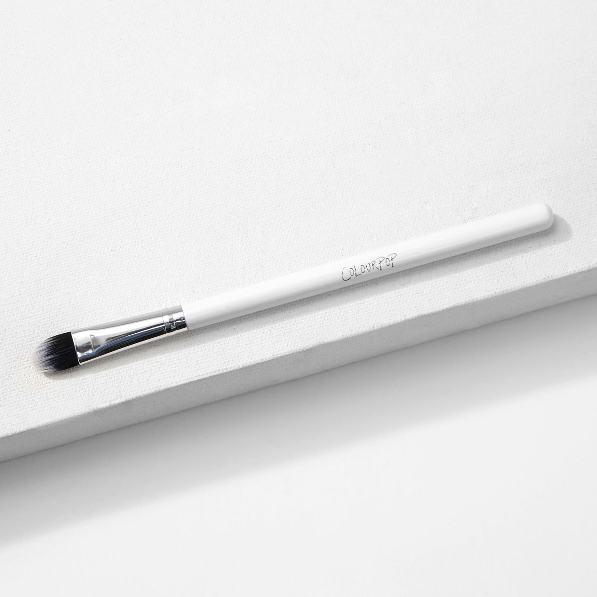 ColourPop Medium Shader Brush with flat oval, dual-toned black-to-white synthetic bristles and a silver ferrule on a white handle, resting on a white textured platform in studio lighting; makeup brush for precise eyeshadow packing and blending.