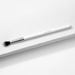 ColourPop Tapered Blending Brush resting diagonally on a white textured surface, featuring a white handle, silver ferrule, and soft tapered black-and-white bristles for seamless eyeshadow blending and crease definition.