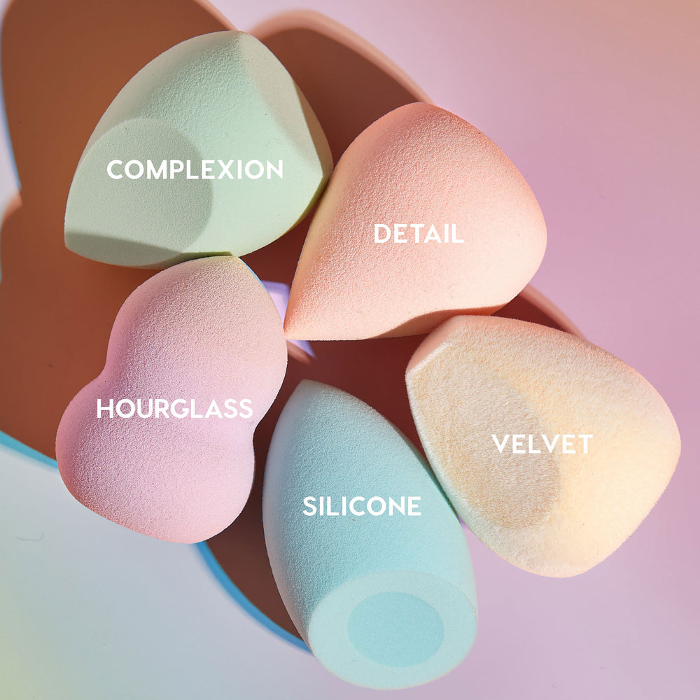 Five pastel makeup sponges from The Full Beat kit fanned out and labeled Complexion, Detail, Hourglass, Silicone, and Velvet, highlighting varied shapes and textures on a soft pink gradient background; ColourPop Makeup Tools & Accessories.