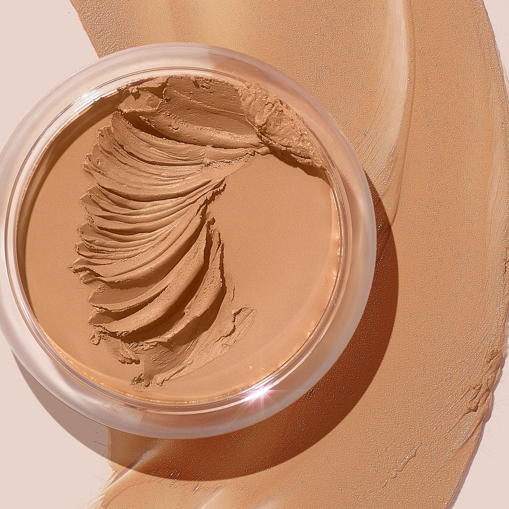 ColourPop SOL Face & Body Bronzing Balm in shade Light sits in a clear round jar with creamy swirls, with warm golden-tan balm swatched around it on a beige surface, showcasing a smooth, blendable texture and natural dewy finish.