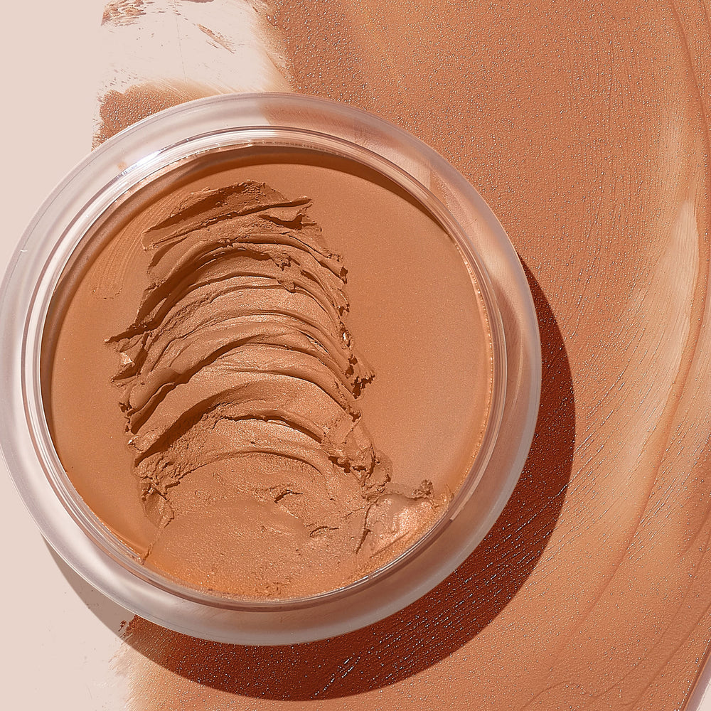 ColourPop SOL Face & Body Bronzing Balm in Medium Dark, creamy bronzer in an open clear jar, shows layered spatula swipes revealing smooth texture, set against broad bronze swatches on a neutral background.