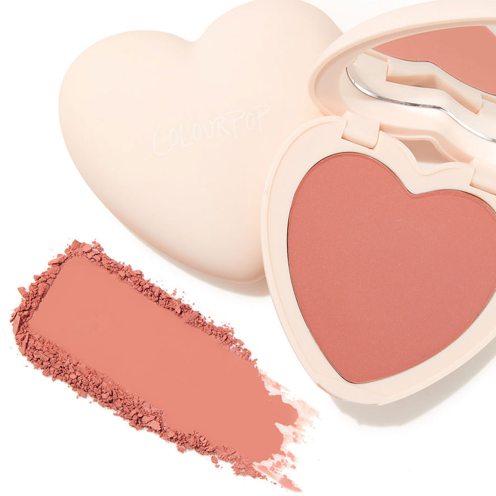 ColourPop Kiss 'N Tell pressed powder blush opens in a cream heart-shaped compact with mirror, revealing a warm peachy-pink heart pan, with a matching crushed swatch beside it on a white background.