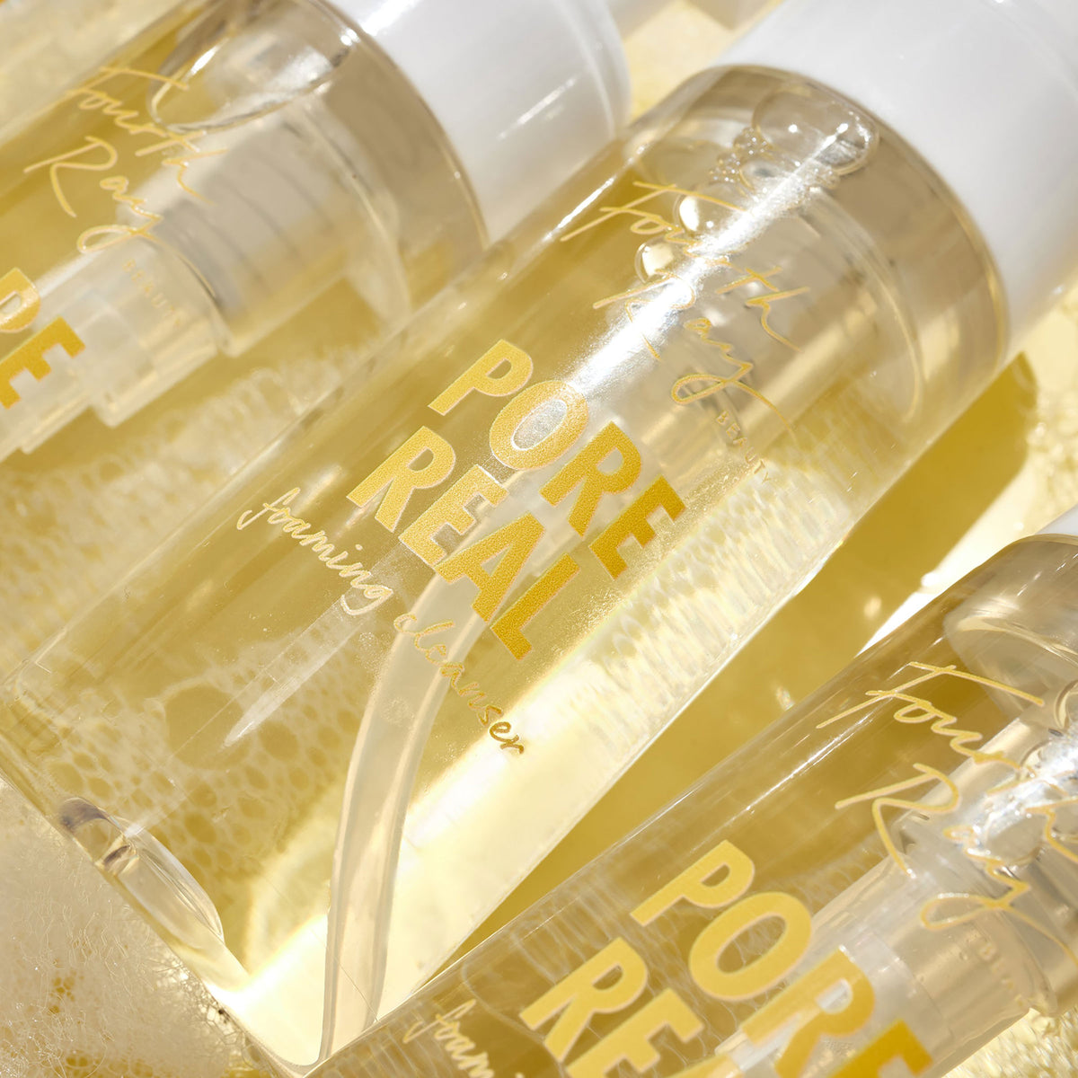 Clear pump bottles of Pore Real Cleanser foaming face wash glistening and tilted on a sudsy, golden surface, white caps and yellow lettering visible; ColourPop Face Cleanser by Fourth Ray Beauty highlighting pore-clearing, gentle lather.
