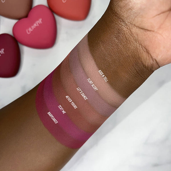 ColourPop Pressed Powder Blush in Text Me swatched on a deep brown forearm, a matte berry-rose stripe among labeled shades (Babycakes, 4ever Yours, Let’s Dance, Flirt Alert, Kiss N Tell), with heart-shaped compacts blurred in the background.