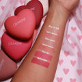 ColourPop Pressed Powder Blush Text Me swatched on a forearm, showing a pigmented rosy pink matte next to Babycakes, 4ever Yours, Flirt Alert, Let’s Dance, and Kiss N Tell, with heart-shaped ColourPop compacts on pink heart-print fabric.