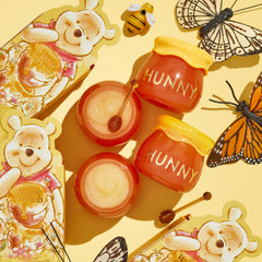 Hunny Pot Lip Care Kit by ColourPop displayed top-down: two honey-pot jars labeled “HUNNY,” open balm and sugar lip scrub with mini honey dippers, Winnie the Pooh themed packaging, bee and monarch butterflies on a yellow background.