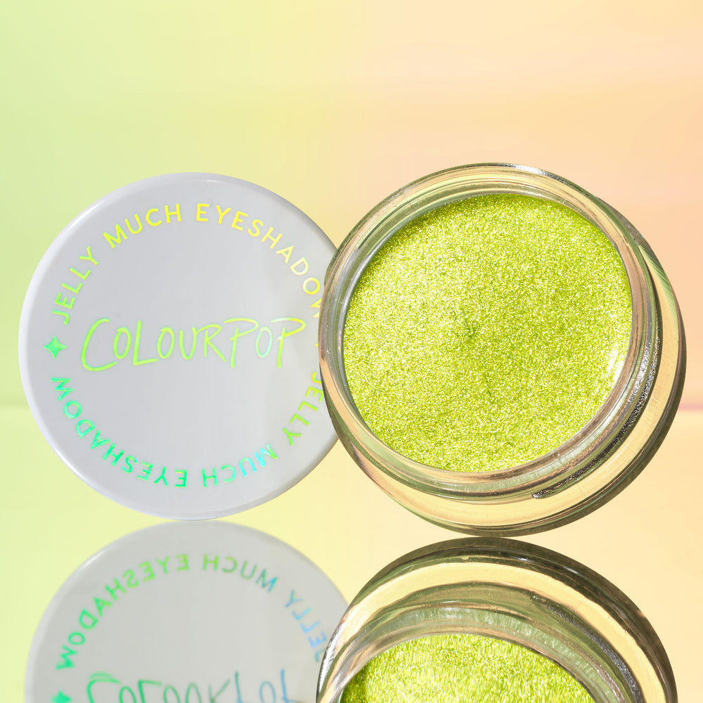 ColourPop Jelly Much Shadow in Billionaire sits open, revealing a metallic lime-green gel shimmer; the white lid with holographic Jelly Much Eyeshadow text rests beside it on a mirrored surface against a soft pastel gradient backdrop.