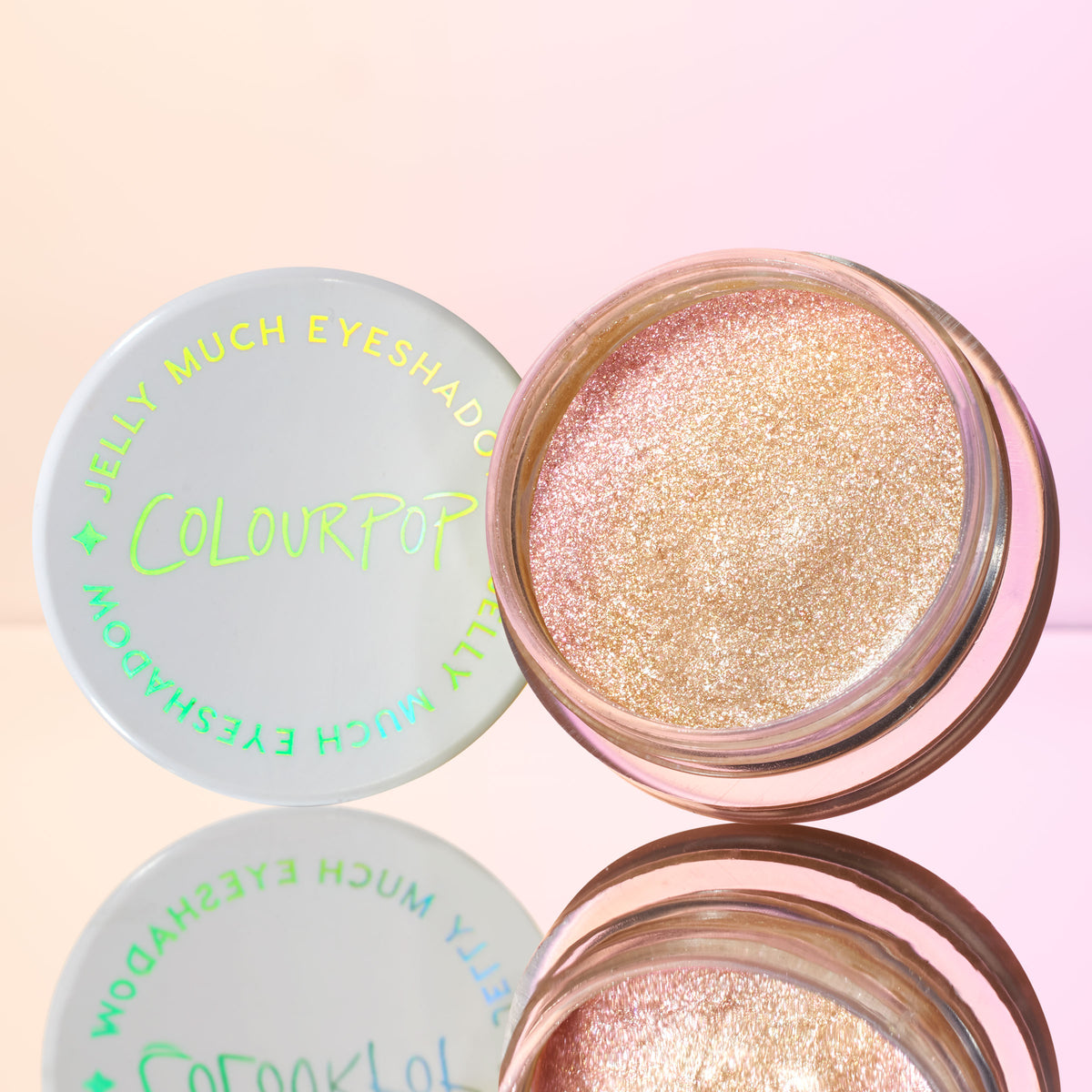 Open pot of ColourPop Jelly Much Shadow, Field of Fairies, revealing champagne‑pink, metallic glitter jelly eyeshadow; white lid with neon green ColourPop logo beside it on a mirrored surface against a soft pink gradient background.