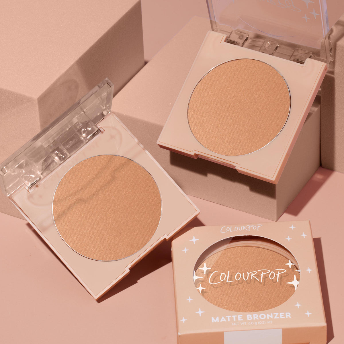 ColourPop Summerland Beach pressed powder bronzer compact sits open to reveal a warm matte tan pan, alongside a second open compact and a boxed unit on blush‑pink blocks; ColourPop Pressed Powder Bronzer for a natural sun‑kissed finish.