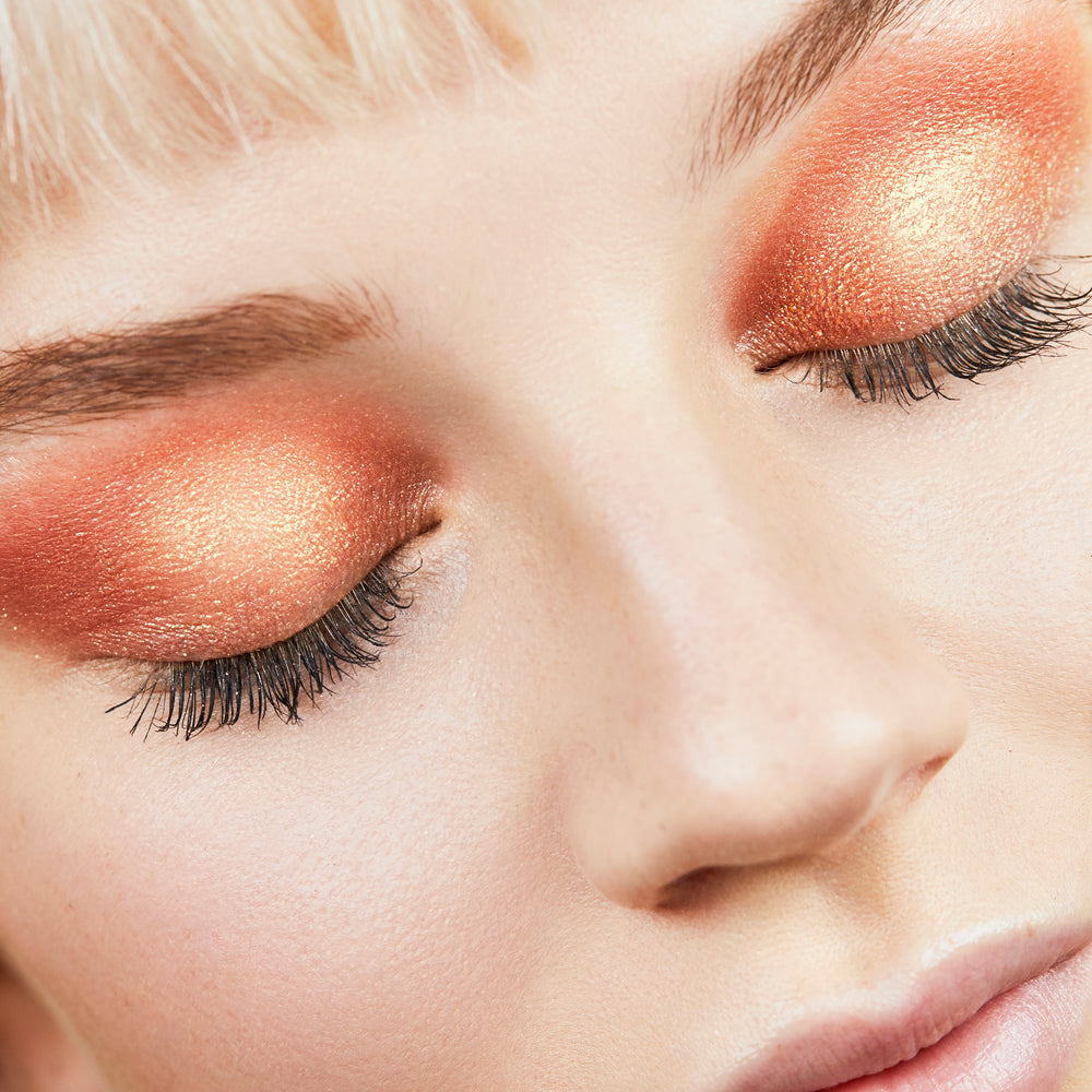 ColourPop Super Shock Shadow in Pledge Class shimmering across closed eyelids, showing a warm copper-to-peach gradient with golden metallic sparkle and soft lashes in a close-up beauty shot.